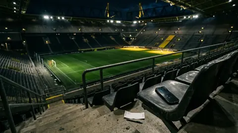 Leeres Fußballstadion im Flutlicht, im Vordergrund liegt ein Smartphone auf einem Tribünensitz